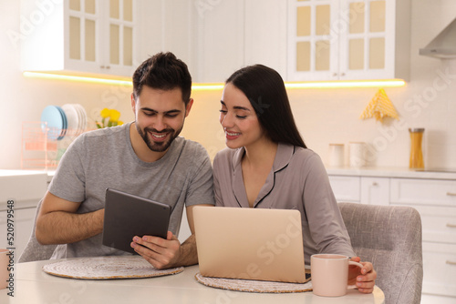 Happy couple in pajamas using gadgets at kitchen table