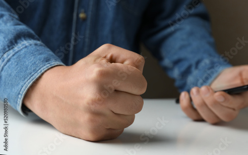 Man with clenched fist holding mobile phone at table, closeup. Restraining anger