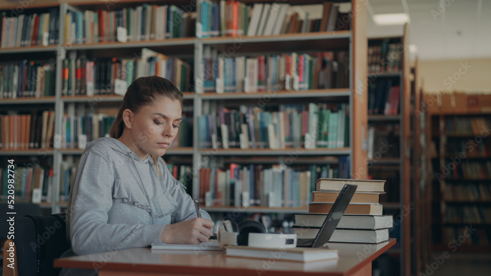 Beautiful female student sitting at table with books in library writing down summary in copybook using laptop computer Beautiful female student sitting at table with books in library writing down summary in copybook using laptop computer
