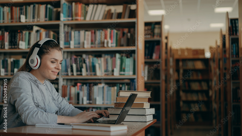 Beautiful positive caucasian female student with big white headphones on head working at table in spacious library in front of laptop. She is smiling and texting Beautiful positive caucasian female student with big white headphones on head working at table in spacious library in front of laptop. She is smiling and texting