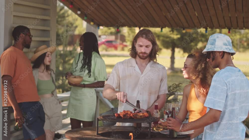 Medium slowmo of group of six young multiethnic friends drinking beer and chatting while having barbecue party at campsite in summer Medium slowmo of group of six young multiethnic friends drinking beer and chatting while having barbecue party at campsite in summer