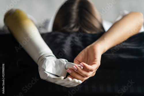 Close up two hands of woman with disability bionic arm on black bed at home, person lost her arm, disabled cyborg girl artificial prosthetic limb, people with special needs 