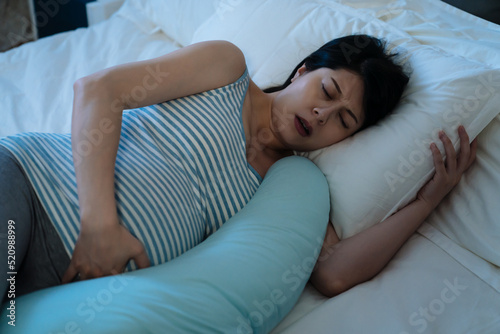 closeup asian woman suffering labor contraction pain is holding her abdomen while sleeping with a pregnancy pillow on bed at midnight in the bedroom. photo