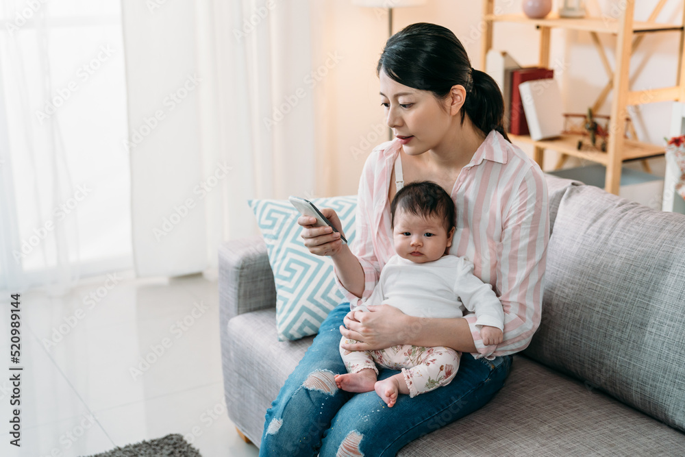 © PR Image Factory - portrait asian mother holding her innocent newborn child in arms is using mobile phone on the sofa in a cozy bright living room at home. © PR Image Factory - portrait asian mother holding her innocent newborn child in arms is using mobile phone on the sofa in a cozy bright living room at home.