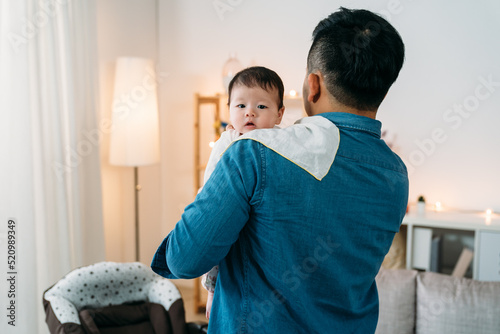Photos view from behind cute curious asian baby is looking at the camera over her dad’s shoulder with burping cloth in a modern cozy home living room at daytime