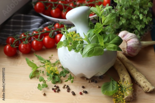 Mortar with fresh herbs near garlic, horseradish roots, black peppercorns and cherry tomatoes on wooden table, closeup