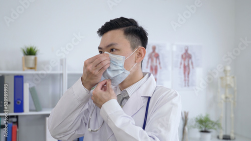 asian male medical worker in doctor’s uniform is making sure to wear the mask properly at a clinic.