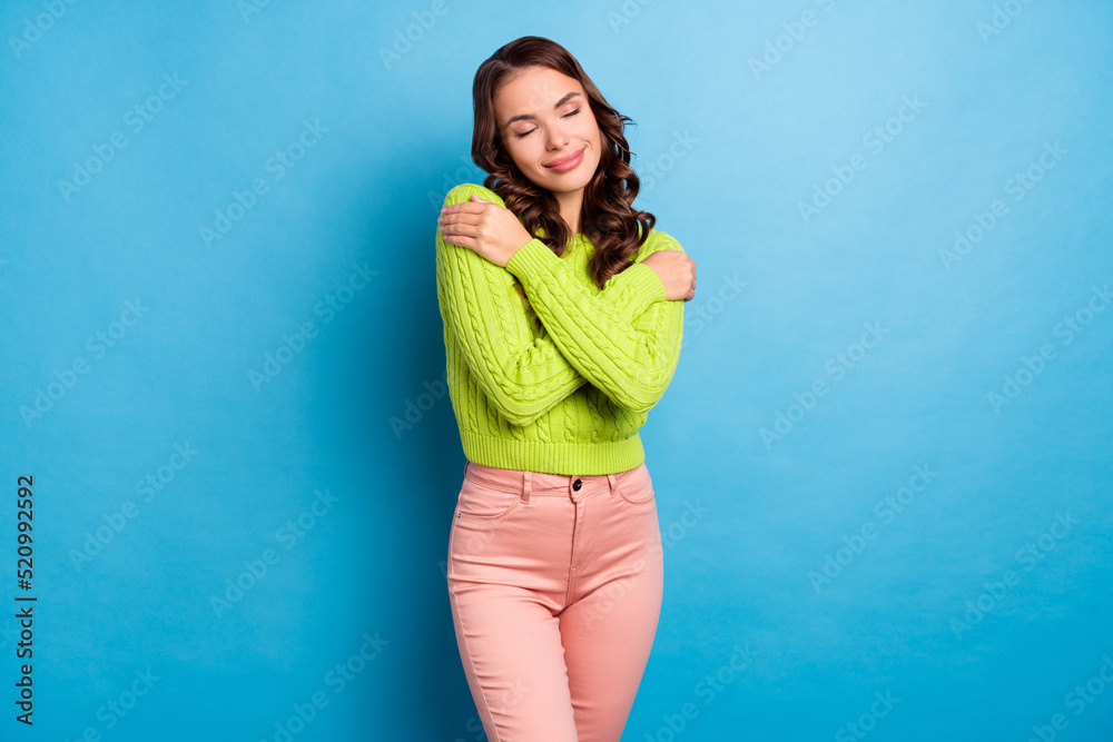 Photo of adorable nice girlish woman with wavy hairdo wear green sweater pink pants hugging herself isolated on blue color background Photo of adorable nice girlish woman with wavy hairdo wear green sweater pink pants hugging herself isolated on blue color background