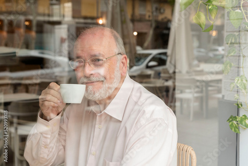 a smiling older man with glasses sitting behind a window in a coffee bar holding a cup of coffee