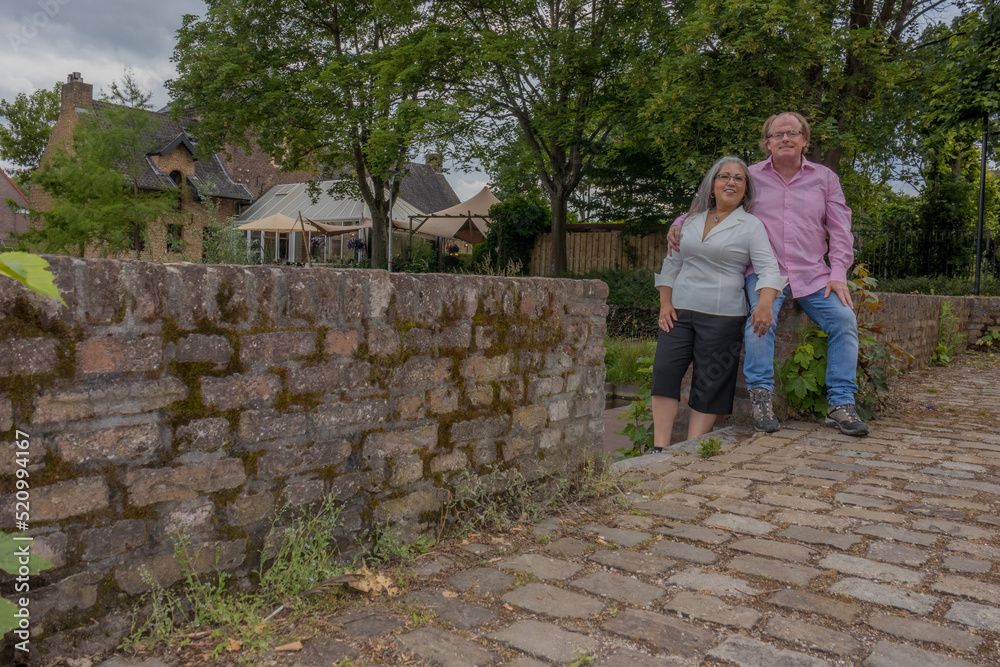 Fototapeta premium Mature couple standing by fence embracing, smiling expression, looking at camera, casual clothing, glasses, relaxed day with trees and houses in background in Heerlen, South Limburg, Netherlands