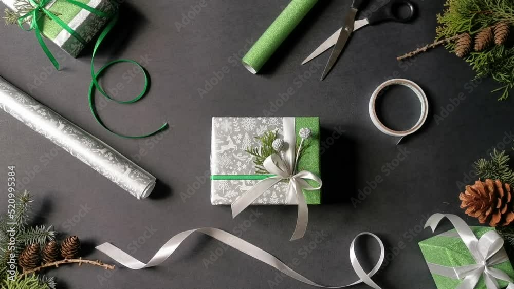 Handmade festive gift-box wrapping. Packaging tools: sharp sciccors, scotch tape, satin ribbons and wrapping paper rolls. Black background with spruce cones, branch of spruce and gift-boxes.