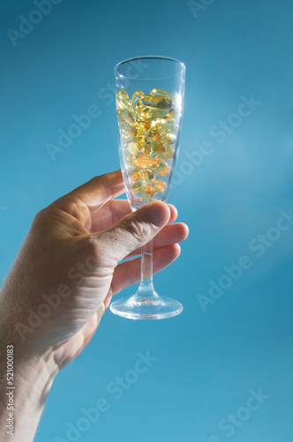 A champagne glass raised in the man's left hand to the light and filled with fish oil pills in front of light blue background. Creative minimal healthy life concept.
