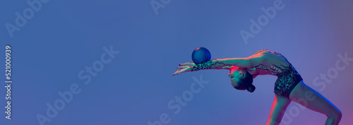 Portrait of young flexible girl, rhythmic gymnast athlete training with blue ball isolated on blue purple background in neon light