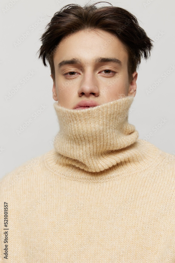 Close-up photo.A cute young man with dark short curly hair combed back in a beige sweater with a high neck stands on a gray background .Vertical studio photo Close-up photo.A cute young man with dark short curly hair combed back in a beige sweater with a high neck stands on a gray background .Vertical studio photo