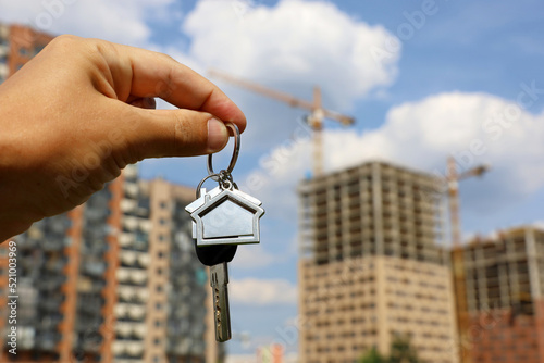 Real estate agent, keychain in shape of a house and key in male hand on background of construction cranes and new buildings. Byung apartment or renting property in summer