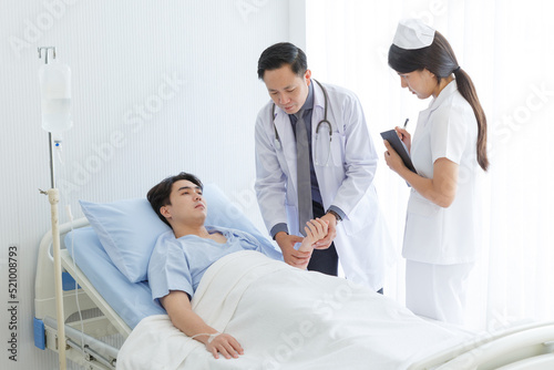 Senior doctor and young male patient who lie on the bed while checking pulse, consult and explain with nurse taking note and supporting in hospital wards.
