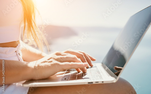 Digital nomad, woman in the hat, a business woman with a laptop sits on the rocks by the sea during sunset, makes a business transaction online from a distance. Freelance, remote work on vacation.