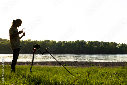 Silhouette of a guy who puts on wireless headphones. In front of him stands a wireless metal detector supported by a shovel. On the river bank, forest in the background