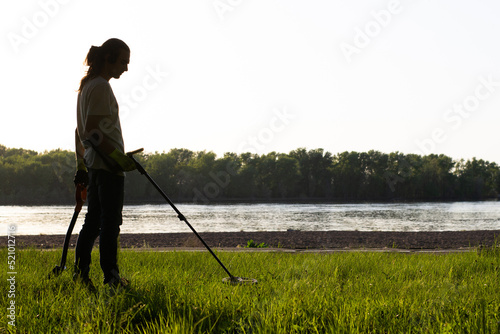 Silhouette of a man with a wireless metal detector on his shoulder and a shovel in his other hand. On the river bank, trees in the background