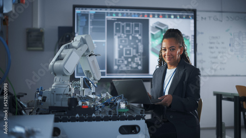 Portrait of Smiling Black Developer Using Laptop while Working with Robotic Arm in Modern University Classroom. Woman Looking at Camera. Success and Programming Concept. Medium Shot