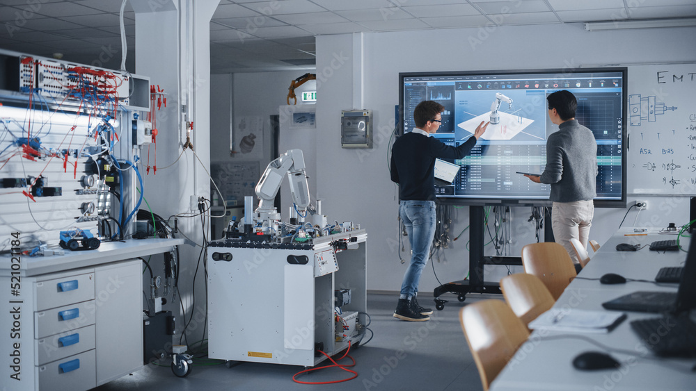 Diverse Team of Two Students Standing Near Big Screen with Tablet and Laptop While Discussing Programming and Technologies. Education and Studying in Modern University of Technology Concept.