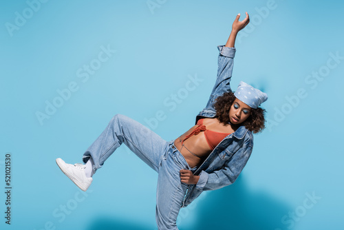 african american woman in denim clothes posing on one leg with arm up on blue background