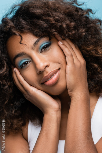 portrait of african american woman with makeup touching face and smiling at camera isolated on blue