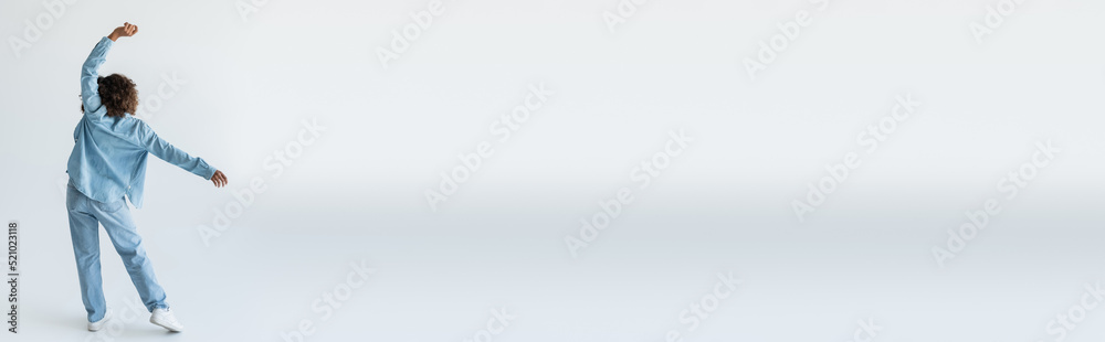 back view of african american woman in denim shirt and jeans standing with raised hand on grey background, banner back view of african american woman in denim shirt and jeans standing with raised hand on grey background, banner
