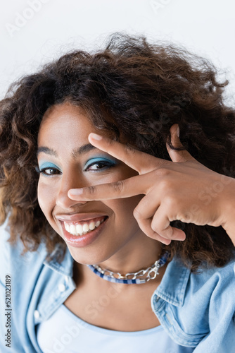 smiling african american woman showing victory sign near face with makeup isolated on white