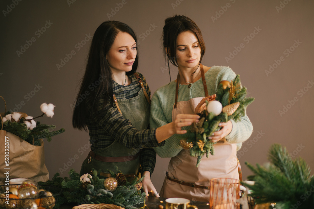 Two millennial women making Christmas wreath using pine branches and festive decorations. Small business