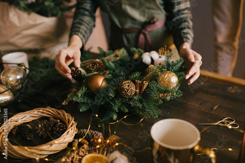 Millennial woman making Christmas wreath using pine branches and festive decorations in workshop. Small business