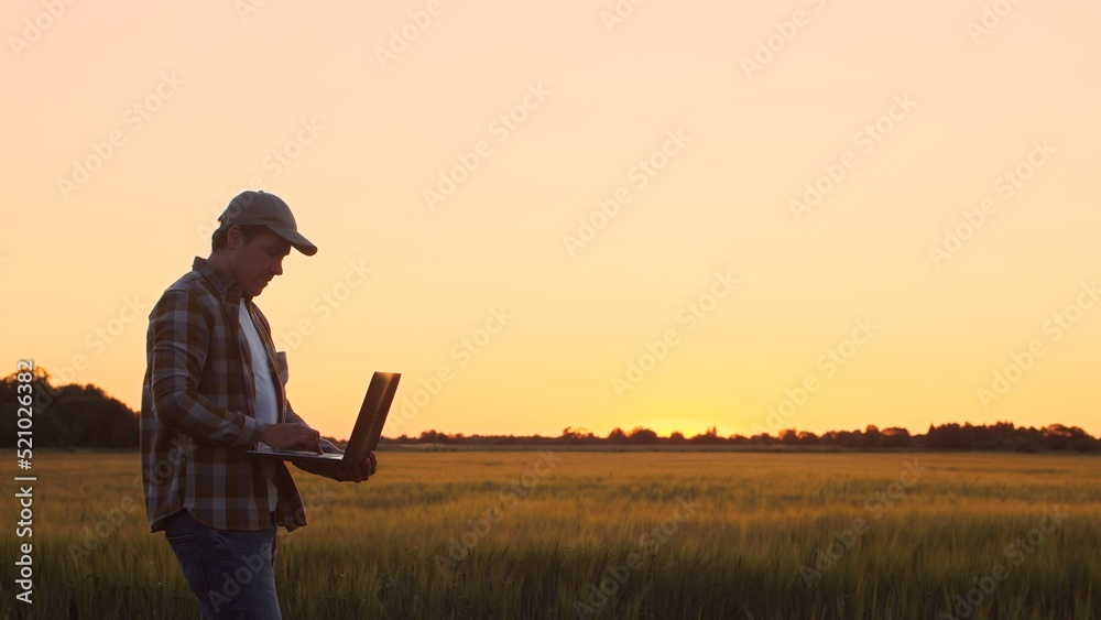 © Acronym - Farmer with a laptop computer in front of a sunset agricultural landscape. Man in a countryside field. Country life, food production, farming and technology concept.