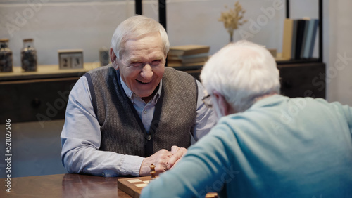 joyful man looking at senior friend while playing chess at home