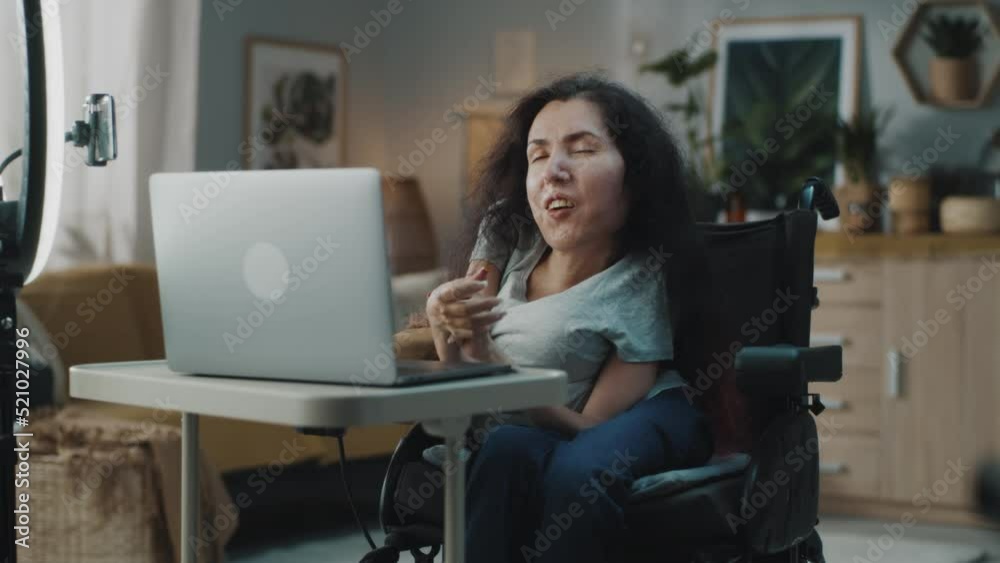 Woman with disability sitting in a wheelchair at home in a cozy room recording a video blog or making a stream in social networks, using a laptop and smartphone on a tripod with a round LED backlight Woman with disability sitting in a wheelchair at home in a cozy room recording a video blog or making a stream in social networks, using a laptop and smartphone on a tripod with a round LED backlight