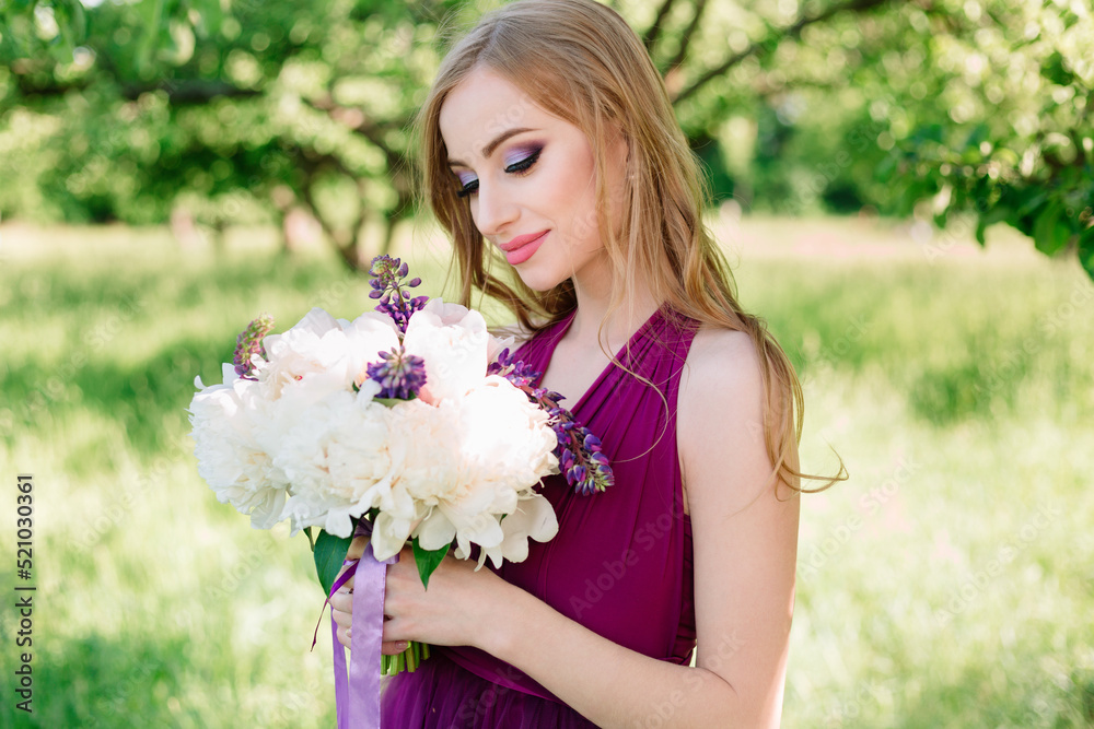 A young attractive woman in a purple dress and with professional makeup poses in an orchard in the summer with a bouquet of peonies in her hands. Bridesmaid at a photo shoot. A young attractive woman in a purple dress and with professional makeup poses in an orchard in the summer with a bouquet of peonies in her hands. Bridesmaid at a photo shoot.