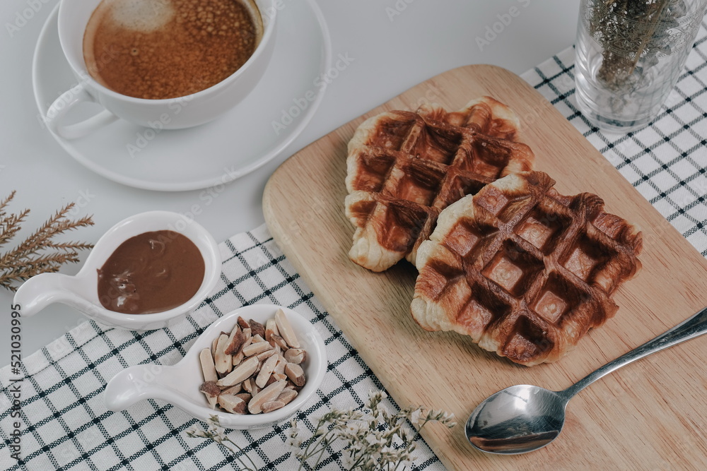 Croffle, Croissant Waffle Korea pastry. Croffle served in wooden plate with almonds, chocolate sauce and coffee on square pattern table cloth. Selective focus. Croffle, Croissant Waffle Korea pastry. Croffle served in wooden plate with almonds, chocolate sauce and coffee on square pattern table cloth. Selective focus.