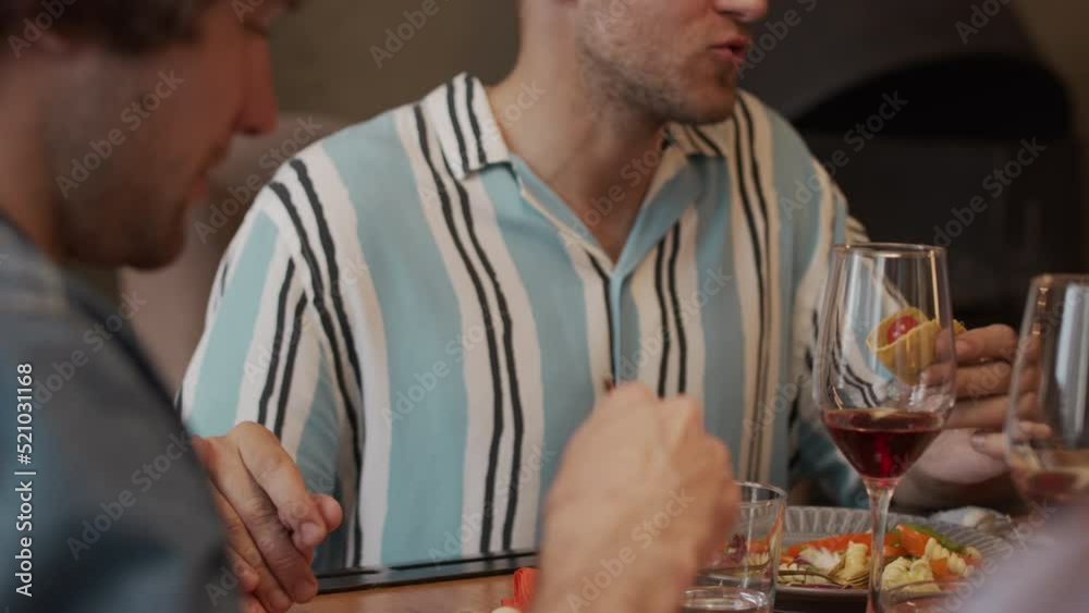Waist up slowmo of happy romantic gay couple sitting at dinner table with friends holding hands and sharing food with each other Waist up slowmo of happy romantic gay couple sitting at dinner table with friends holding hands and sharing food with each other