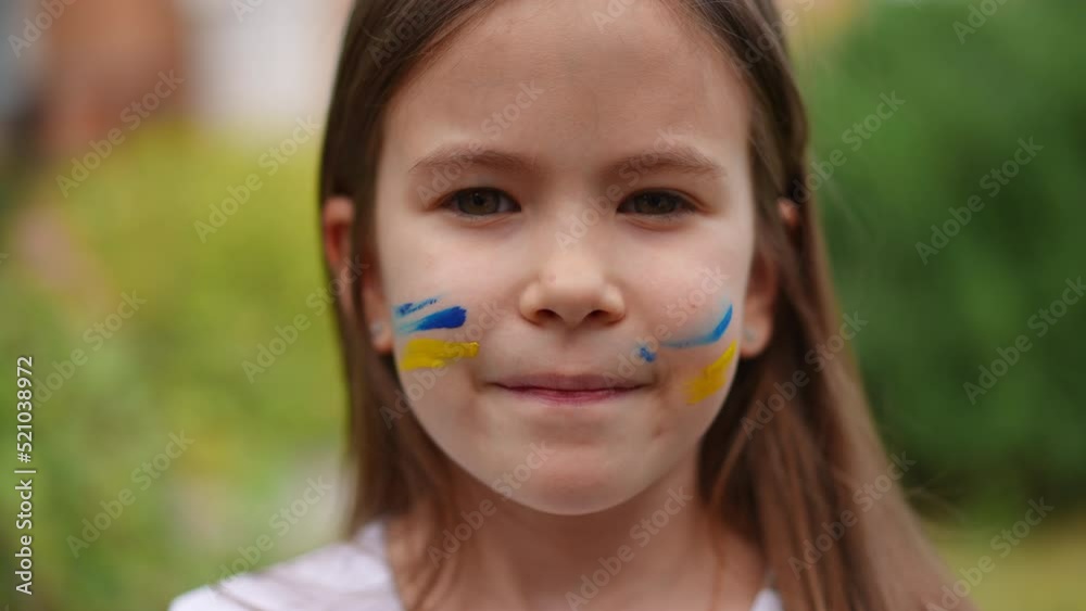 Pretty girl with brown eyes and face painting looking at camera smiling. Ukrainian child with flag colors on cheeks posing in slow motion outdoors. Pride and childhood concept Pretty girl with brown eyes and face painting looking at camera smiling. Ukrainian child with flag colors on cheeks posing in slow motion outdoors. Pride and childhood concept
