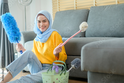 Slika na platnu Young happy muslim woman wearing yellow gloves  and holding a basket of cleaning supplies in living room