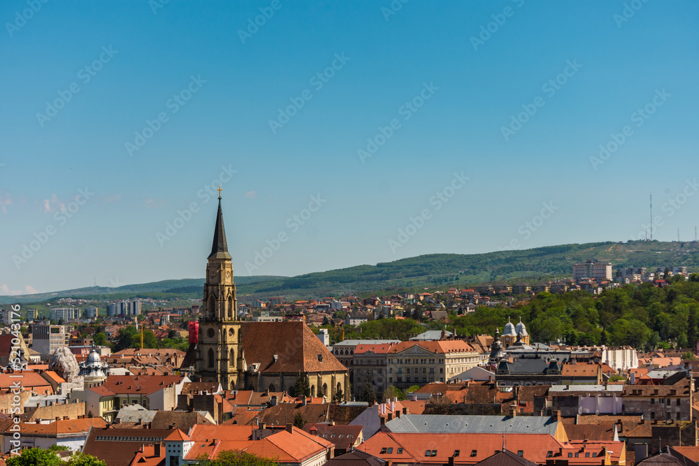 Cityscape of Cluj Napoca with church and vintage buildings on clear blue sky. Aerial view of an old town with hills in the distance