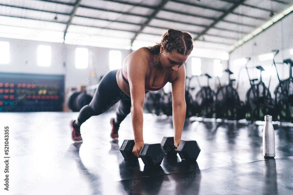 Strong female in tracksuit doing plank on hands for training strength of muscles, slim fit girl exercising push ups with dumbbells during cardio workout in gym studio reaching body positive