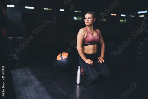 Contemplative Caucasian fitness girl resting with sportive ball and water thermos for refreshing after hardwork training in gym studio, thoughtful female athlete thinking about cardio workout