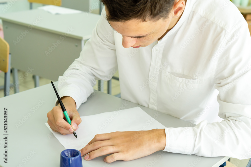 A student writes with a fountain pen while studying at school, sitting at a desk. Portrait of a college guy writing with a pen while completing a task. High quality photo A student writes with a fountain pen while studying at school, sitting at a desk. Portrait of a college guy writing with a pen while completing a task. High quality photo