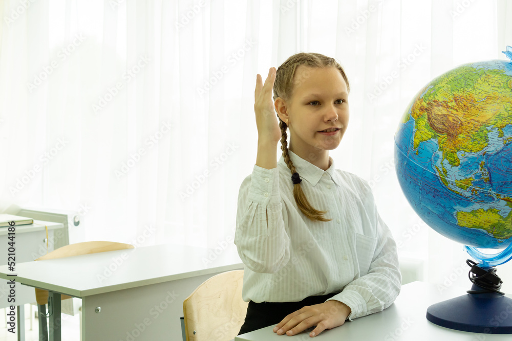 A smiling student giving an answer in class with her hand raised is sitting at a school desk in a white shirt. There is a globe on the table. The concept of a geography lesson. High quality photo A smiling student giving an answer in class with her hand raised is sitting at a school desk in a white shirt. There is a globe on the table. The concept of a geography lesson. High quality photo