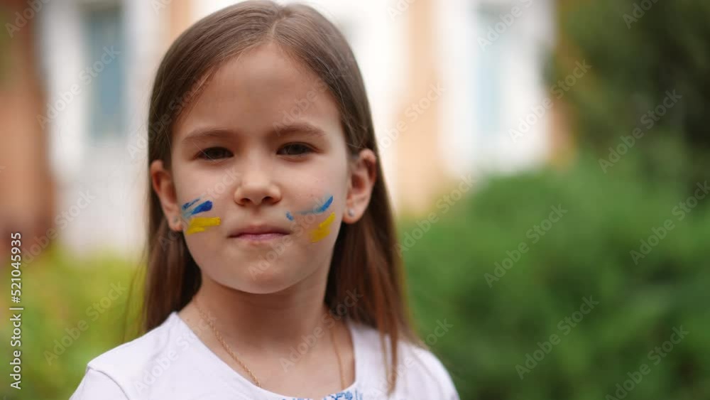 Close-up Ukrainian girl with serious facial expression turning looking at camera in slow motion. Portrait of pretty child posing outdoors with national flag colors face painting Close-up Ukrainian girl with serious facial expression turning looking at camera in slow motion. Portrait of pretty child posing outdoors with national flag colors face painting
