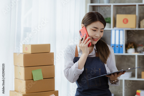 Asian woman is talking on the phone with a customer to confirm an order, she owns an online store, she packs and ships through a private transport company. Online selling and online shopping concepts.