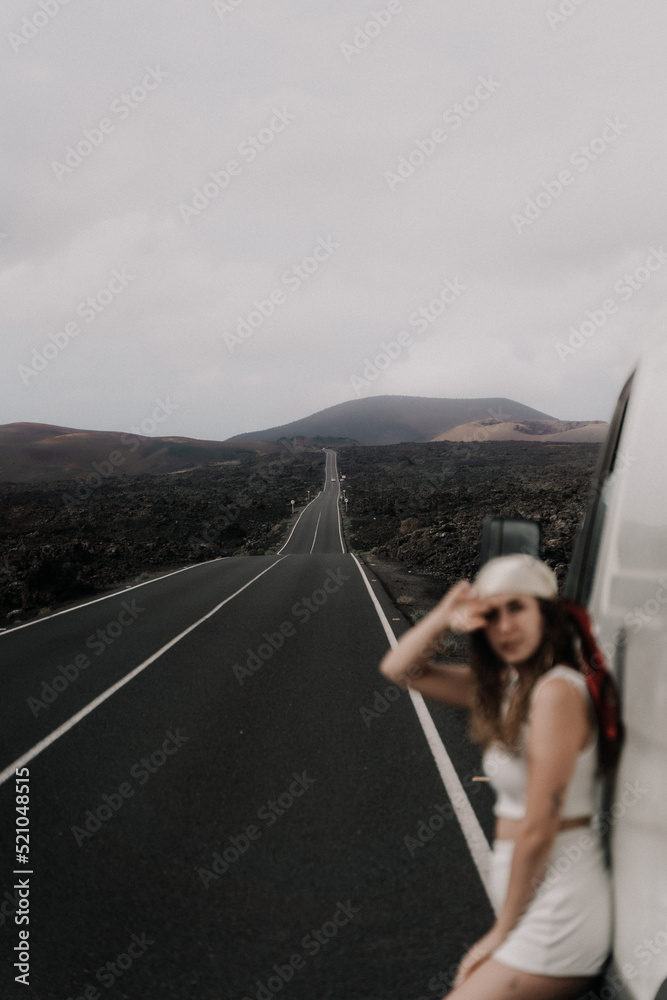 joven chica posando sobre su furgoneta con un bonito paisaje volcánico en Lanzarote y una carretera recta