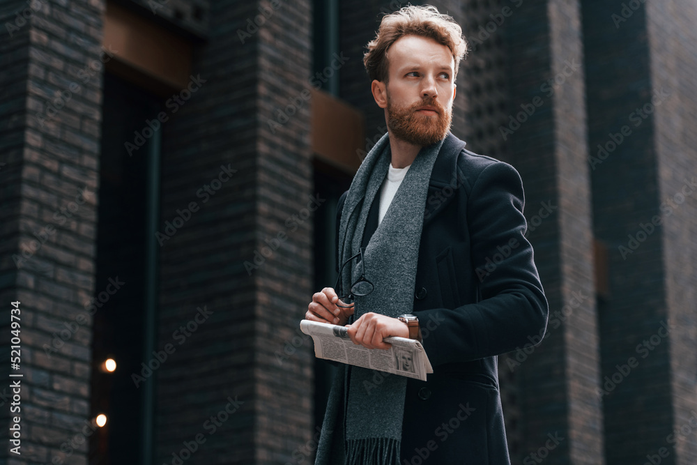 Fototapeta premium Holding newspaper in the hand. Stylish man with beard is outdoors near building