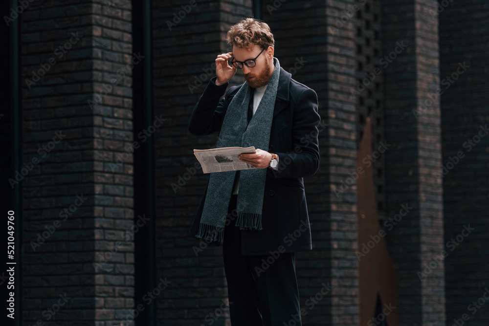 With newspaper in hands. Stylish man with beard and in glasses is outdoors near building With newspaper in hands. Stylish man with beard and in glasses is outdoors near building