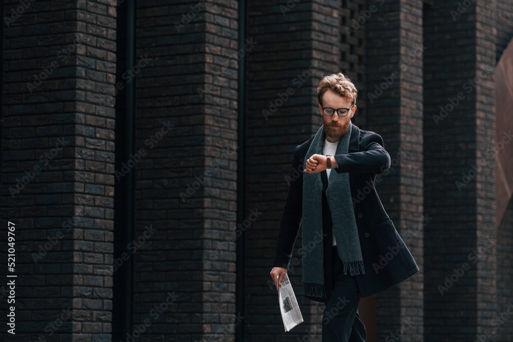Checking time. Stylish man with beard and in glasses is outdoors near building Checking time. Stylish man with beard and in glasses is outdoors near building